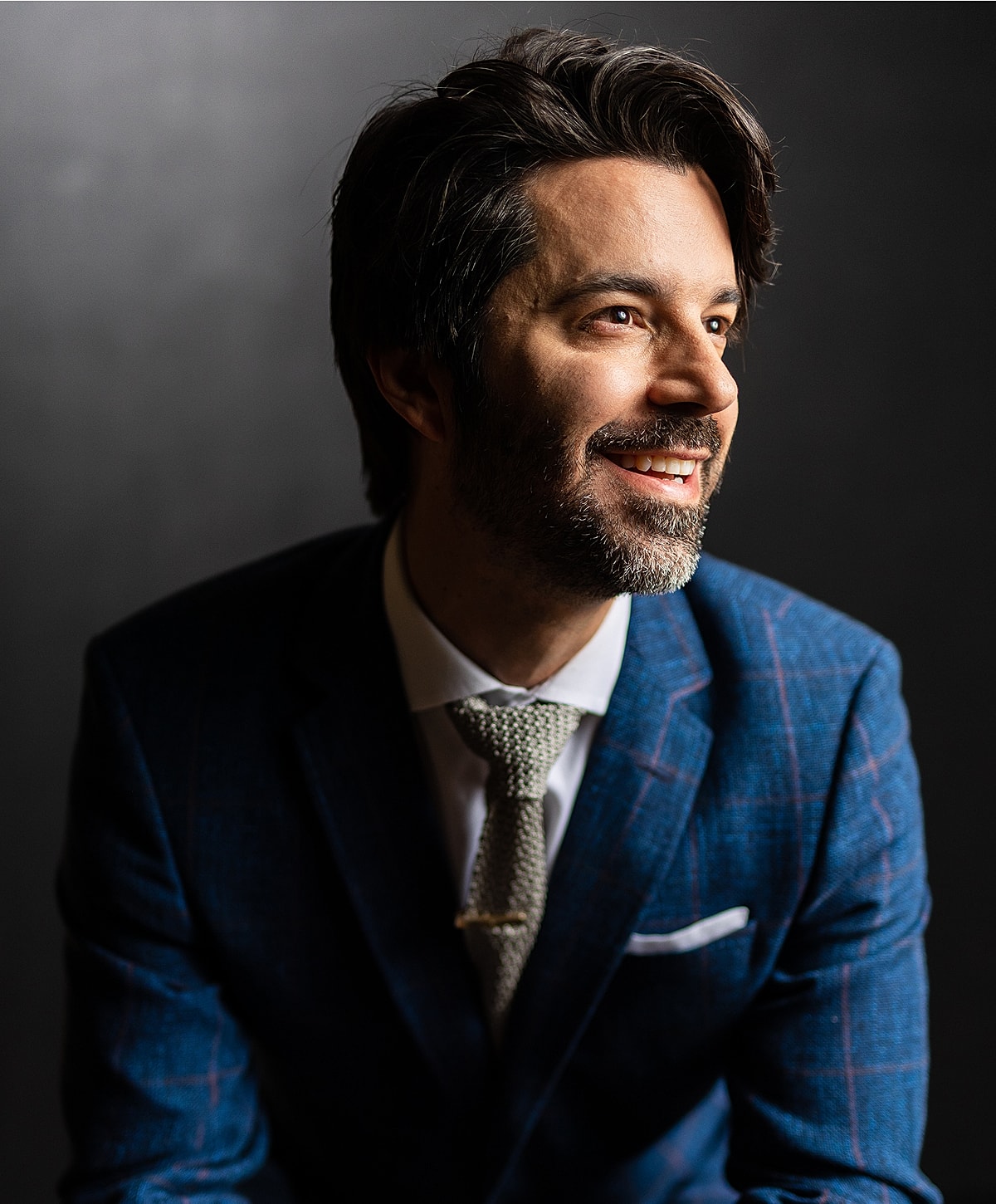 Man in a suit smiling against dark background.