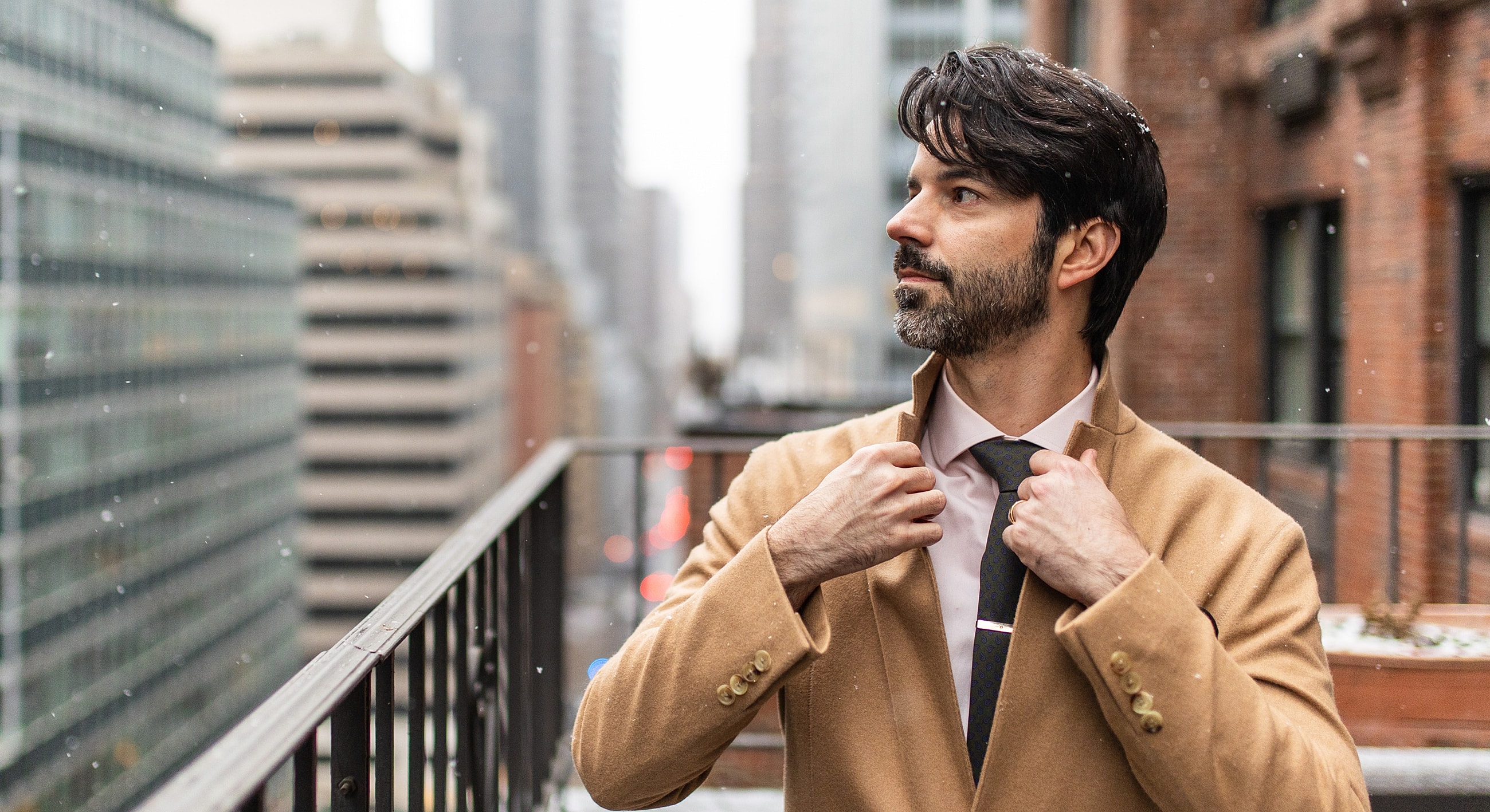 Man adjusting coat in city during snowfall.